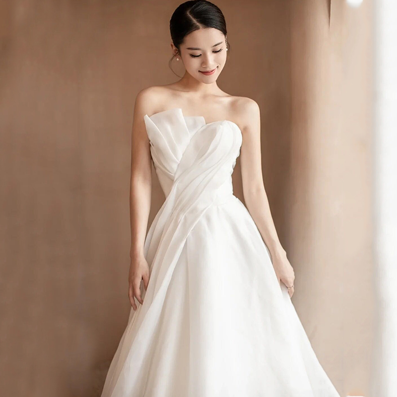 A full-body wedding photo captures the elegance of a bride wearing a MIRACLANDS modern and simple tube top wedding dress. The minimalist design, paired with three-dimensional slim tailoring, beautifully highlights the smooth lines of the waist, creating a light and soft silhouette. This MIRACLANDS Chinese wedding dress seamlessly blends Eastern and European styles, making it an ideal choice for those seeking modern wedding attire. Perfect for the modern bride, this fusion wedding dress is easily accessible through wedding gowns from China online, showcasing the beauty of women dressed from China in a dress perfect for a ceremony wedding.